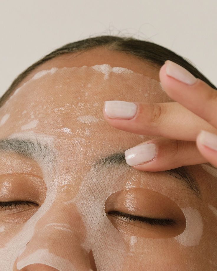 A hand applying a hydrating sheet mask to the forehead