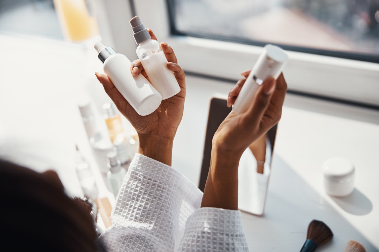 Hands holding several skincare bottles over a crowded vanity
